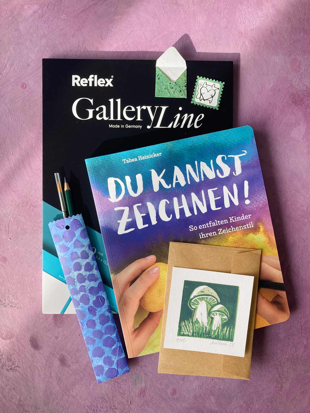 Herbstverlosung 2025: Unser zweites Bündel. Schön aufgestapel im weichen Sonnenlicht, liegen die Preise auf Altrosa. Pinsel und Bleistift gucken aus einer schön gepunkteten Papiertüte.