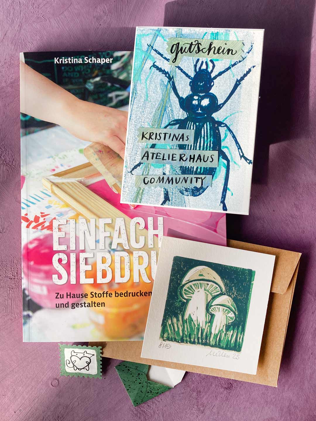 Das letzte Bündel für die Herbstverlosung 2025: in Blau bedruckter Gutschein, gestaltet von Michaela mit Kalligrafie von Tabea liegt direkt auf dem Buch von Kristina.