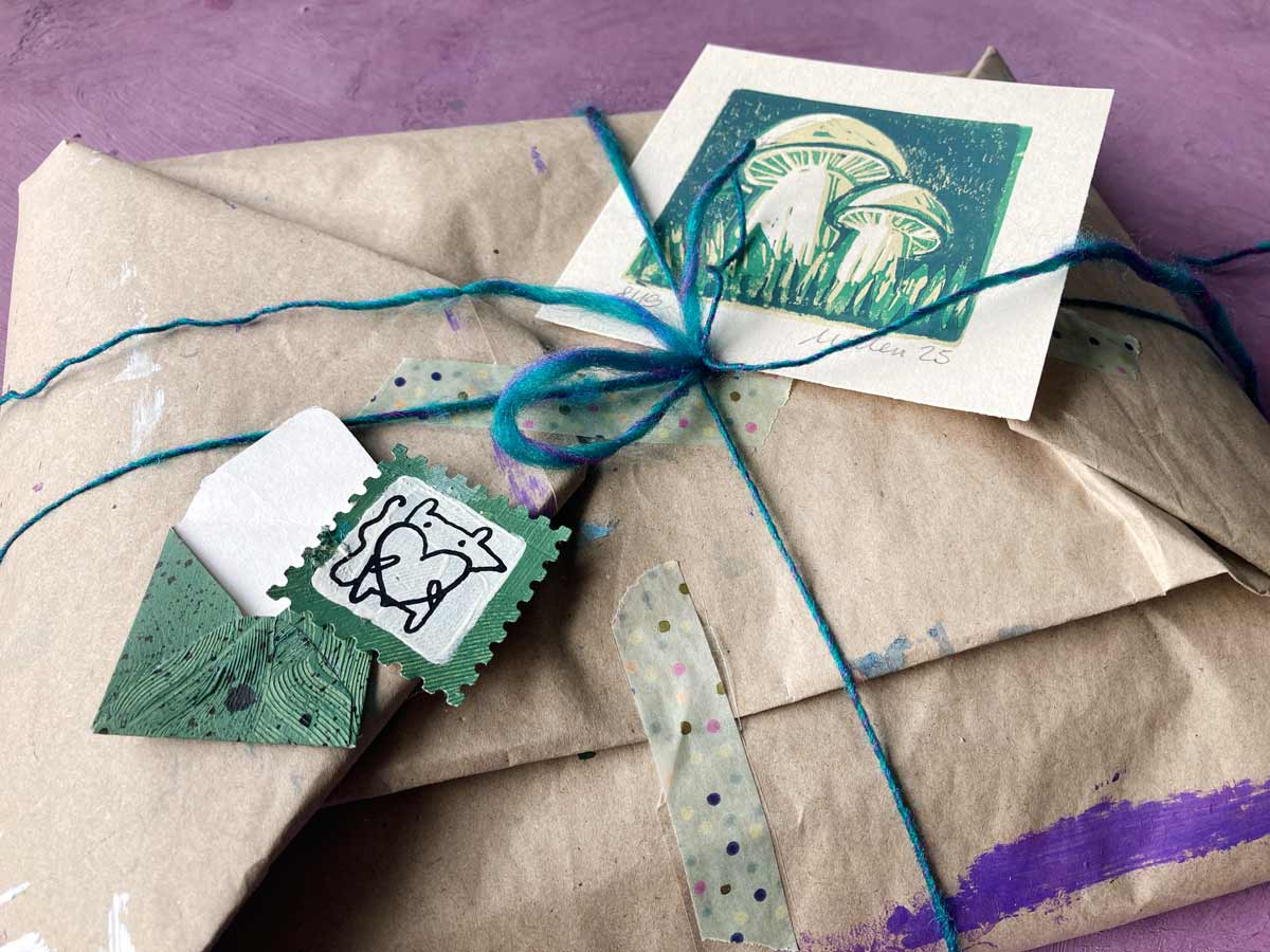 Herbstverlosung 2025 im Postkunstwerk - auf dem Foto siehst du ein wunderschön verpacktes Geschenk, garniert mit zwei kleinen Kunstwerken von Michaela (Pilze grünlich im Reduktionsdruck und Mausita auch grünlich von Tabea)